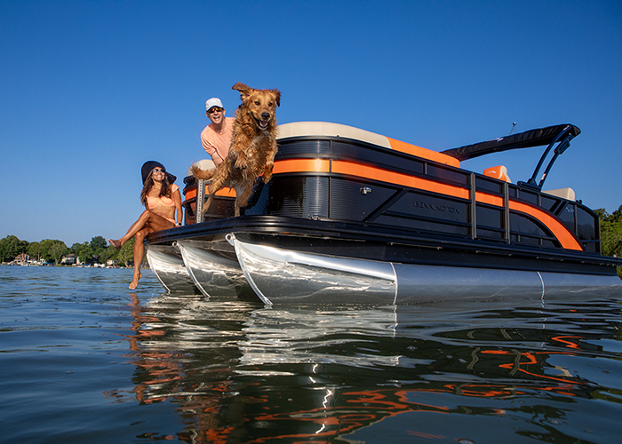 Dog jumping off a black and orange pontoon boat into the water while an adult male and female look on.
