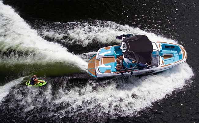 Overhead view of a blue 2018 Super Air Nautique G23 wakeboard boat with a wakesurfer behind the boat on a bright yellow surfboard.