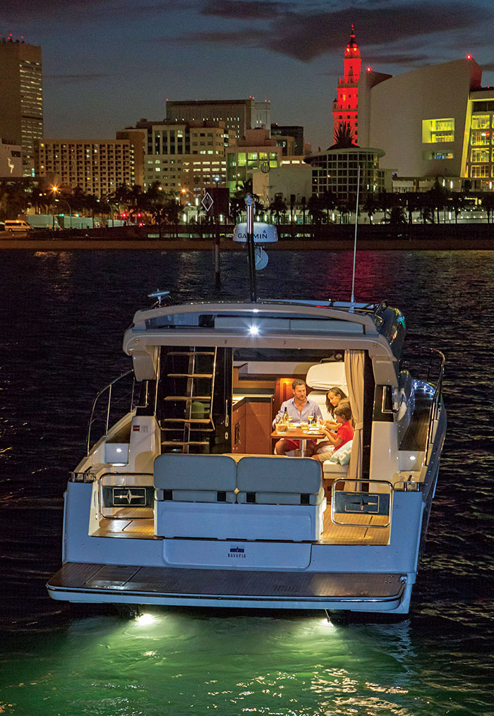 Photo from behind boat at night with a family eating dinner inside the boat and a city in the background