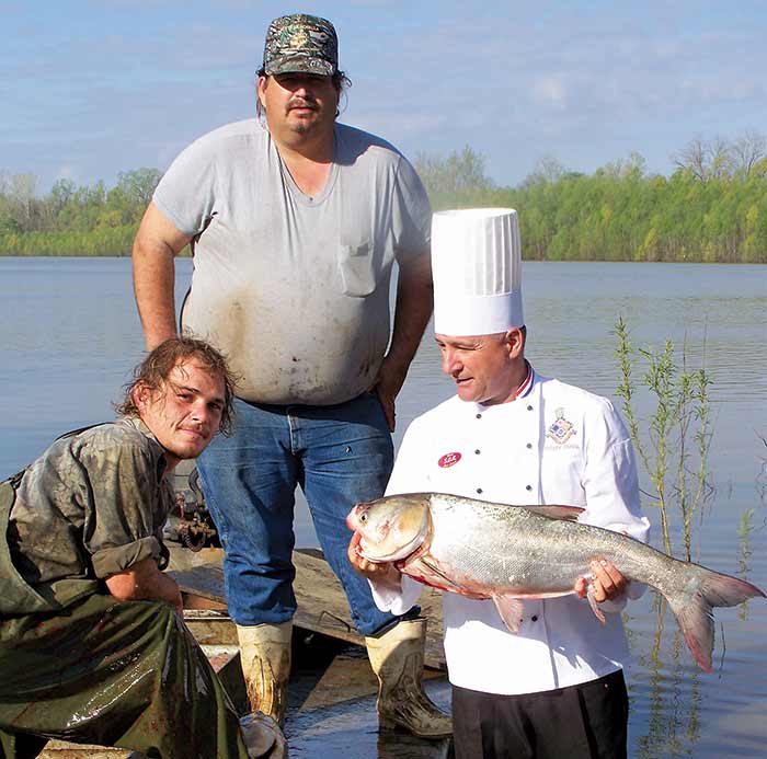 Two men wearing camoflauge and boots covered in red fish blood pose on a flats boat next to a man standing in the water wearing a chefs hat and shirt holding a bloodied fish