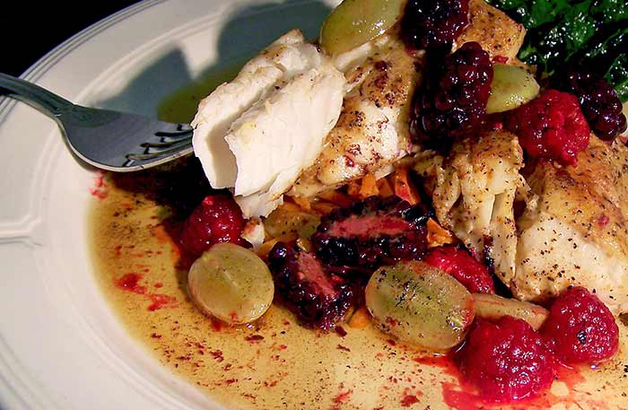 A white plate of food, with white  fish covered in a mix of blackberries, raspberries and grapes