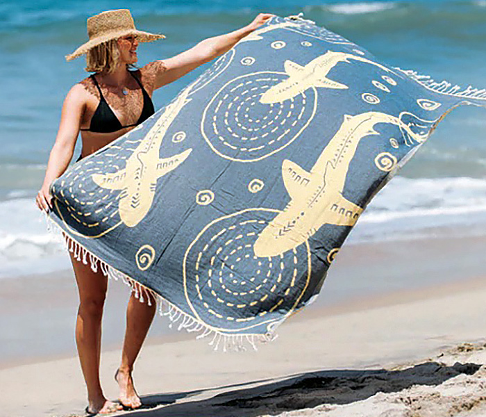 Female wearing a tan hat, black bikini on the beach holding a large blue and white towell.