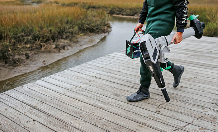 Adult in a green wet suit carrying a battery and a Newport NT300 on a wooden dock.