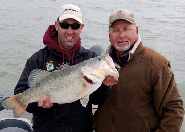 Two white men dressed for cold winter weather smile for the camera, one man holds up a large bass fish.
