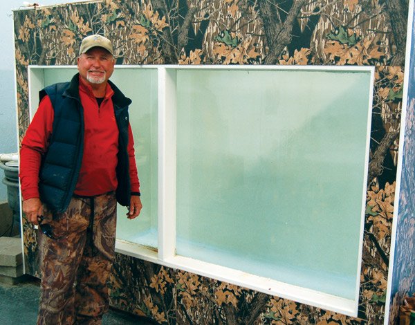 A white man with a red shirt and blue vest stands against a camo wall with two empty glass panes.