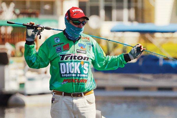 man in green fishing tournament clothes and red had is standing with both hands holding a fishing rod horizontally across his back shoulders