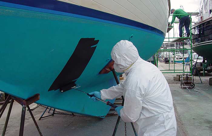 Man wearing full body paint suit with head covering and dust mask painting bottom of a boat using both hands to roller brush on black paint