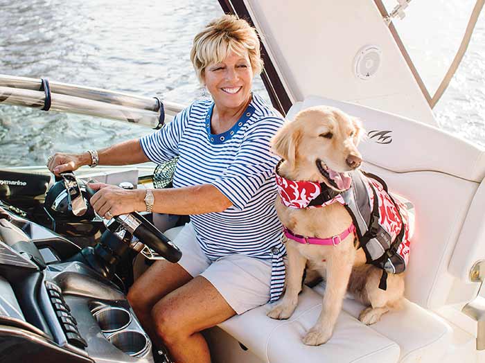 A woman with short blonde hair at the helm of a boat with a golden retriever in a red lifejacket sitting next to her on the white bench seat.