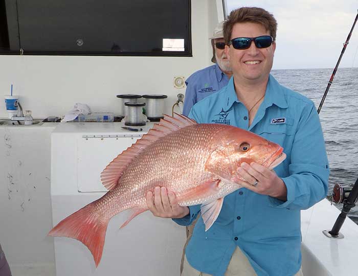 A man in a blue shirt holds a red snapper fish aboard a boat