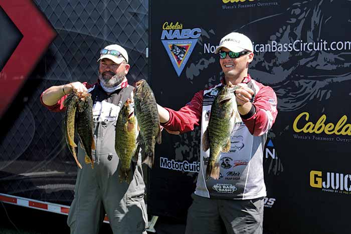 Two men in bass fishing uniform shirts hold up a total of 5 fish 