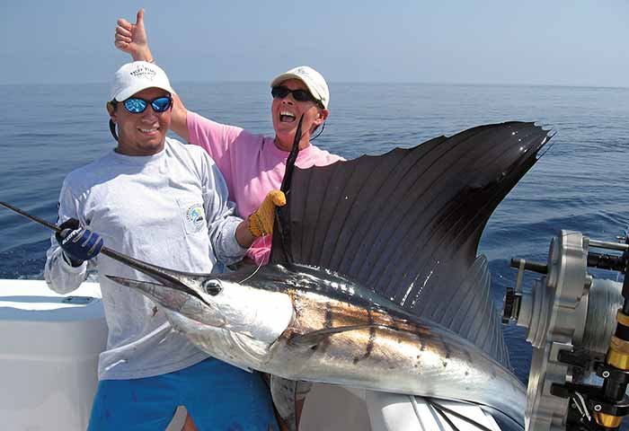 A man holds a billfish with a woman behind him giving an enthusiastic thumbs up 