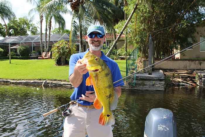 A man in a blue hat and sunglasses smiles holding up a yellow and green bass fish