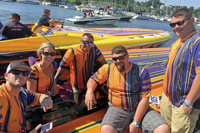 A group of four men and one woman in matching orange and blue shirts smile at the camera from a orange and blue raceboat