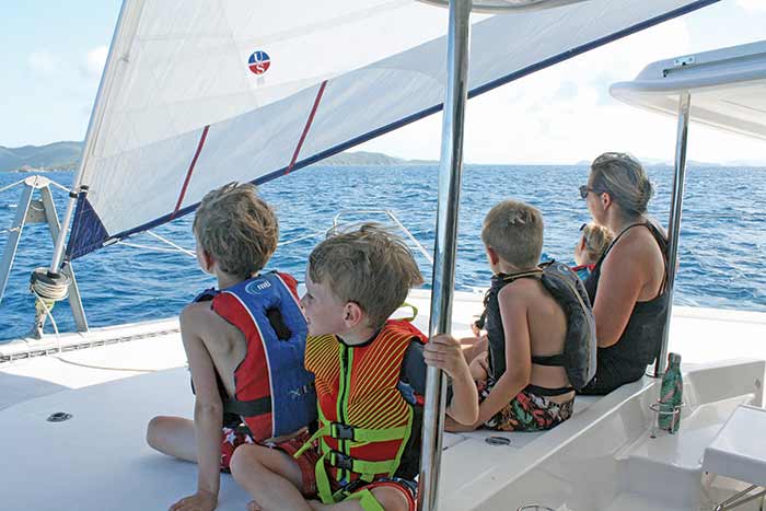 A woman and four kids all in lifejackets stare out into the distance at the water from aboard a catamaran boat
