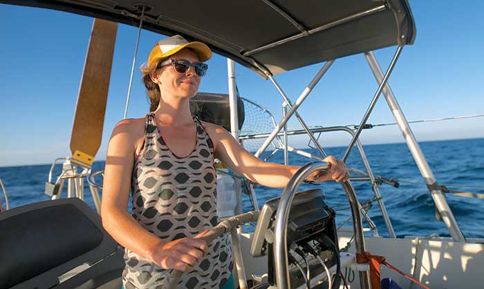 A woman wearing a baseball hat and sunglasses at the helm of a sailboat