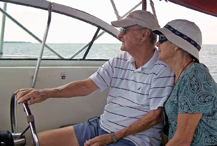 An elderly man and women look out to the horizon from the helm of a boat, in which the man has his hand on the wheel