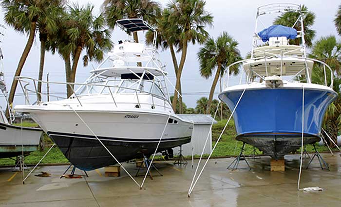 A white sport fishing boat and a bright blue boat are both on metal stands tied to the ground with palm trees in the landscape background