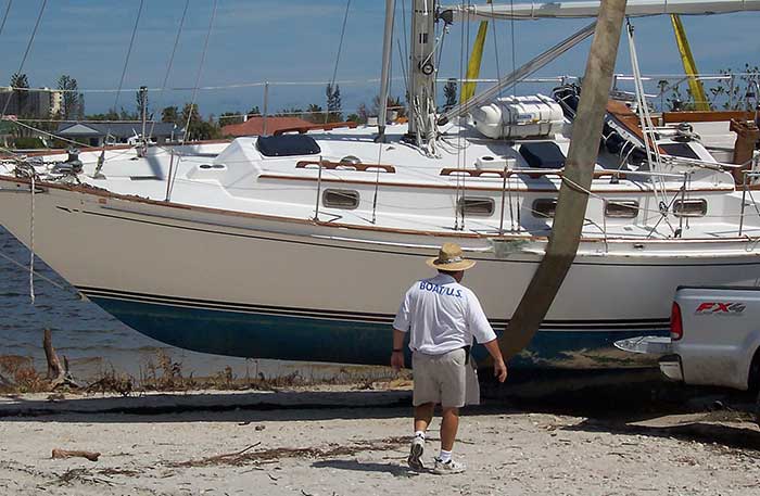 A man with the words BoatUS across the back of his shirt walks toward a damaged blue and white sailboat held in a large sling on land