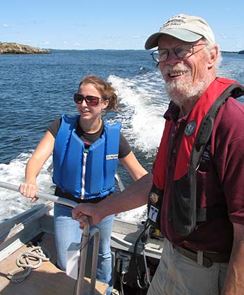 An older gentleman in a red lifejacket and a woman in a blue lifejacket both hold on to a silver bar aboard a boat underway