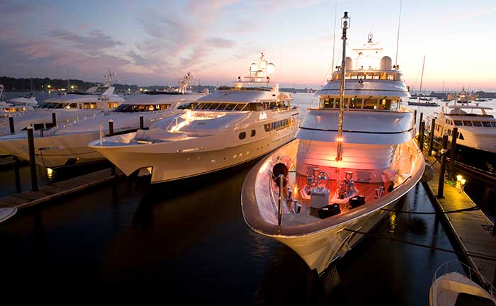 Three large powerboats docked at a marina as the sun sets, one boat has interior lights on