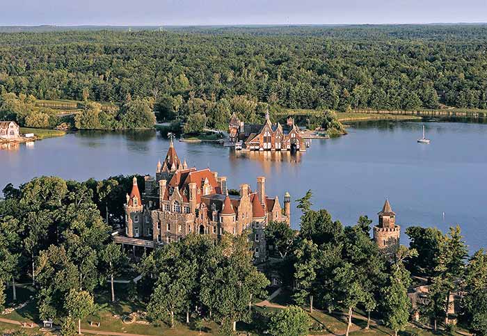 Aerial photo of castle-like brick buildings nested within green trees on a body of water.