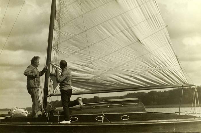 Old sepia photo of Albert Einsein standing on a sailboat rigging the sails with other man helping him