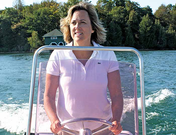 Woman wearing white polo shirt looking off center of camera at helm of powerboat with two hands on steering wheel. 