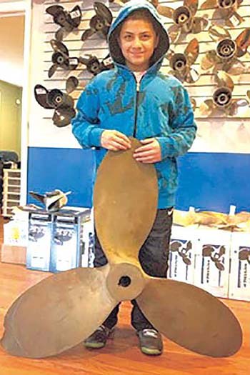 Young boy wearing a blue hoddie standing and holding a boat propeller in front of him