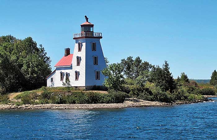 A white lighthouse with several windows and a red roof is centered in the photo, surrounded by a rocky shoreline and green trees and bushes