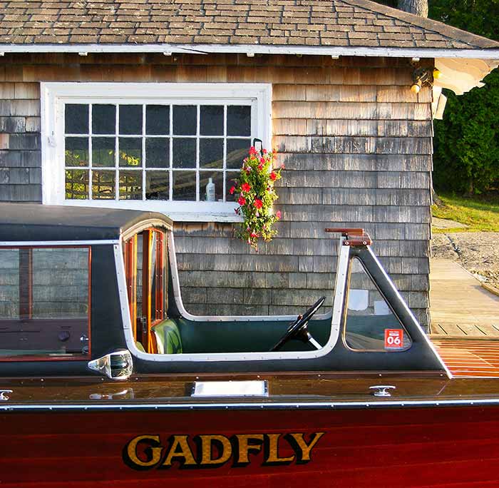 Old wooden boat with name "GADFLY" in gold lettering next to a wooden building with white window panels.