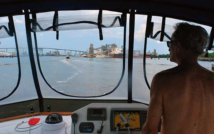 Mna standing at the helm of a large powerboat looking out the cockpit windows with a bridge and city buildings in the background