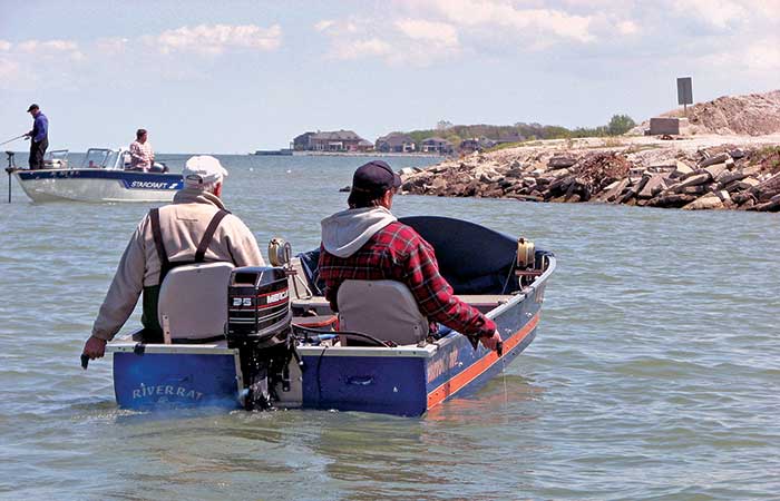 Two men sitting in a small fishing boat heading to a small outcrop of land with one man holding a fishing line in the water