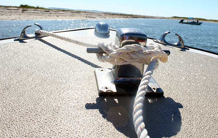 Close-up photo of a line tied to a cleat of a boat with water , a beach and another small boat in the background
