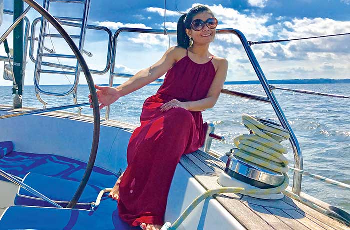Woman sitting on the eedge of a sailboat wearing a long red dress, sunglasses and long daggly earrings holding the boats stering wheel