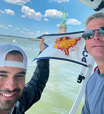 Man wearing a backward facing white ball cap holding a flag with another man wearing sunglasses sailing through the water with the Statue of Liberty in the background