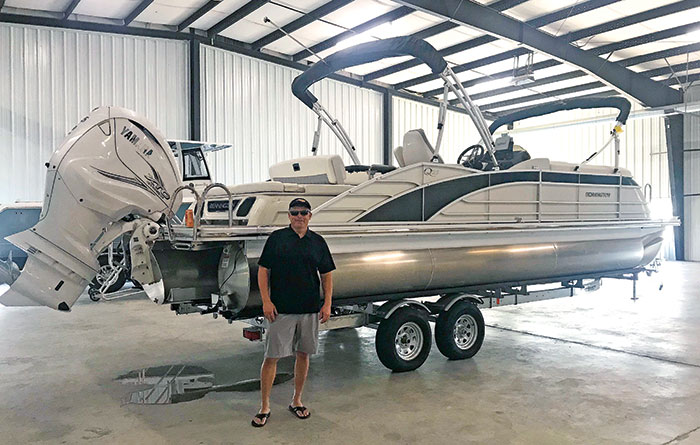 Man wearing a black polo shirt, gray shorts, black ball cap and sunglasses standing in front of white and chrome pontoon boat on a boat trailer in a large hangar