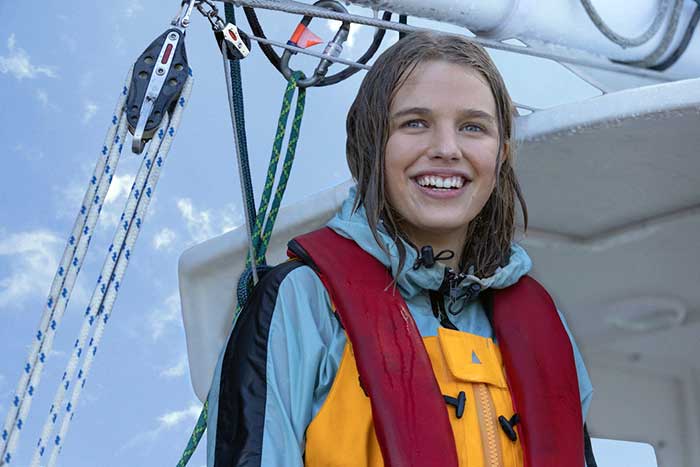 Close-up photo of a girl wearing a red lifejacket, light blue jacket and yellow waders, soaking wet, standing on a sailboat