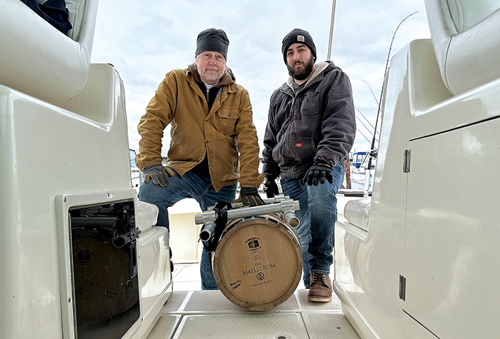 Two men kneeling on the deck of a white boat next to a barrel of whiskey.
