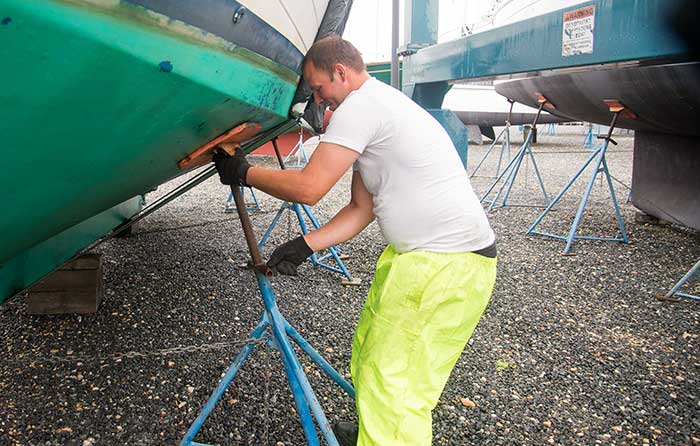 Man wear yellow work pants and a white t-shirt securing a jackstand under the hull of a large boat