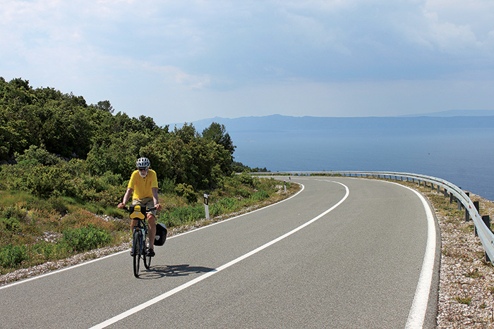 A senior adult male wearing a white helmet and yellow shirt riding a bike on a road