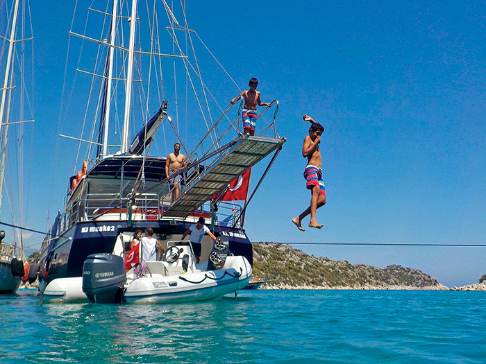Family jumping off a plank and swimming from a large blue and white sailboat on a sunny day