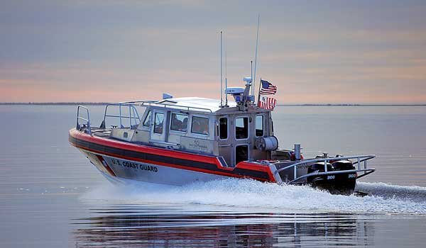 A silver boat with US Coast Guard  labeled on the side cuts across open glassy water, with white spray coming from behind the boat's 2 engines.
