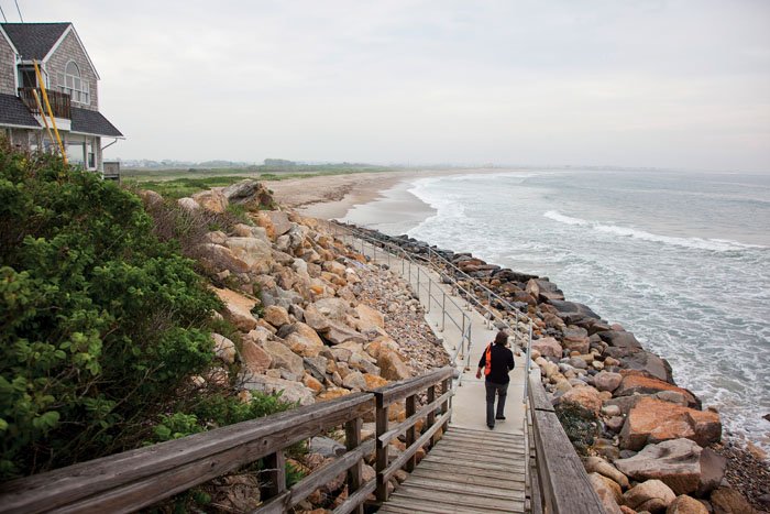 A person wearing black walks down wooden steps as the photograph captures the rocky shoreline and waves crashing on one side of the stairs with more rocks and greenery and a building on the other side.