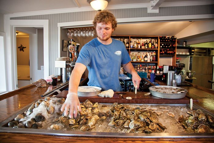 A man in a tshirt is pulling oysters from a icy cooler, and arranging the oysters on smaller icy round trays. A well-stocked bar is in the background of the photo.