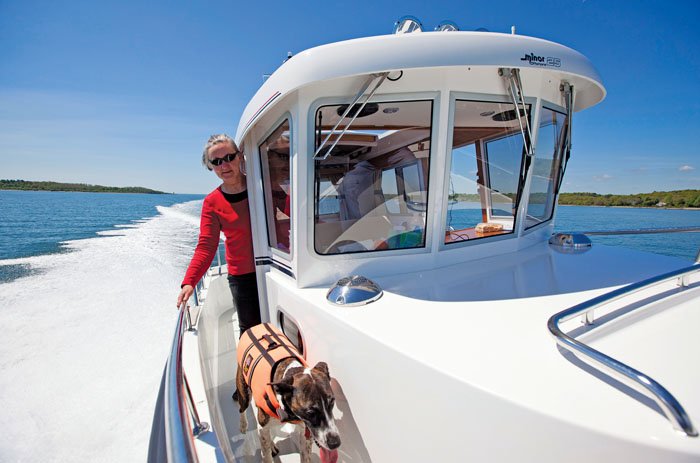 A brown and white dog wearing an orange lifejacket walks toward the front of the white powerboat as it's underway. A woman in a red shirt holds on to the silver rail of the boat and watches the dog move forward. A white wake is seen behind the boat.