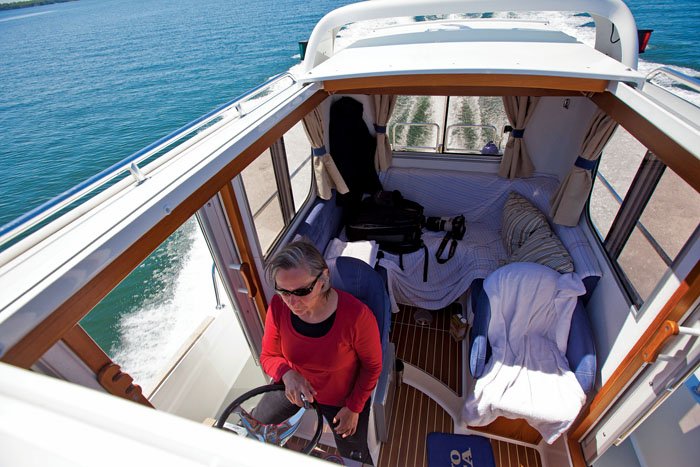 A grey-haired woman wearing sunglasses is at the helm of a white powerboat, you can view the white wake behind the boat through the pilothouse windows.