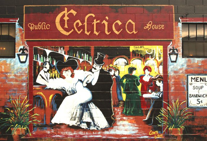 A painted mural of a pub featuring women in fancy dresses and a man in a top hat with the words Public Celtica House as a sign at the top of the mural. 
